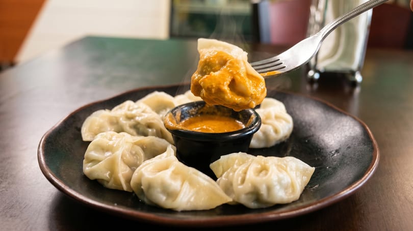 A fork lifts a steaming momo dumpling dipped in orange sauce from a black plate with more dumplings.