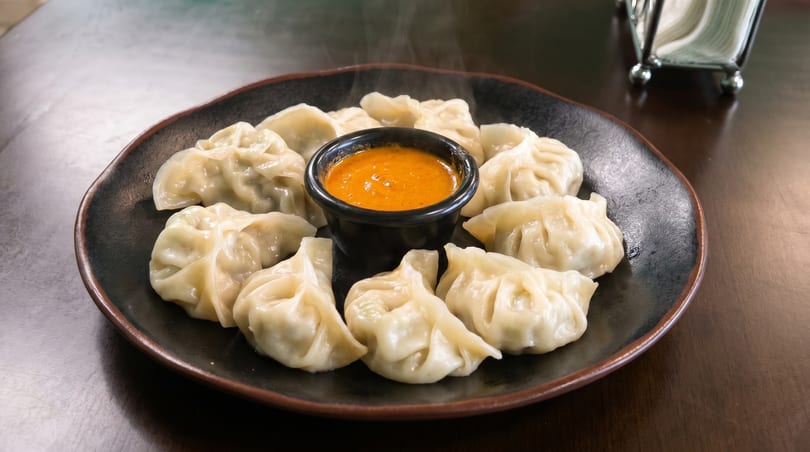 Steaming momos, a type of dumpling, arranged on a dark plate with a small bowl of orange sauce.