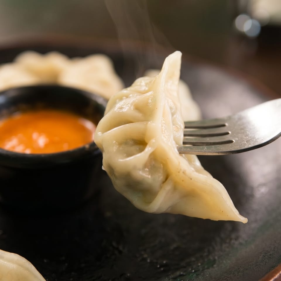 Steaming momo dumpling on a fork, with more dumplings and orange dipping sauce on a dark plate.