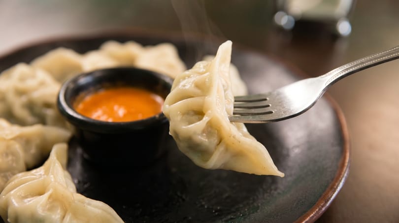 Steaming momo dumpling on a fork, with more dumplings and orange dipping sauce on a dark plate.