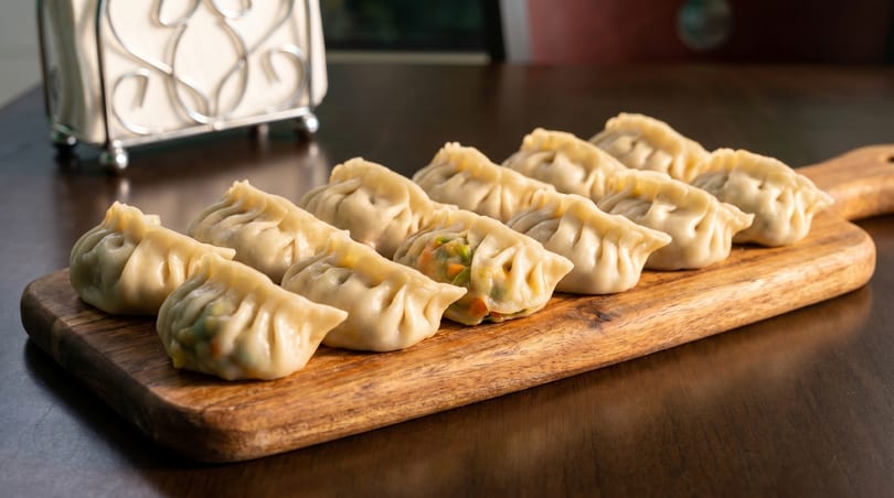 A row of steamed vegetable dumplings, with one showing its colorful filling, on a wooden board.