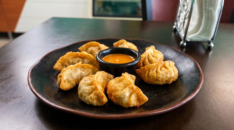 Golden-fried momos arranged around a small bowl of orange dipping sauce on a dark plate.