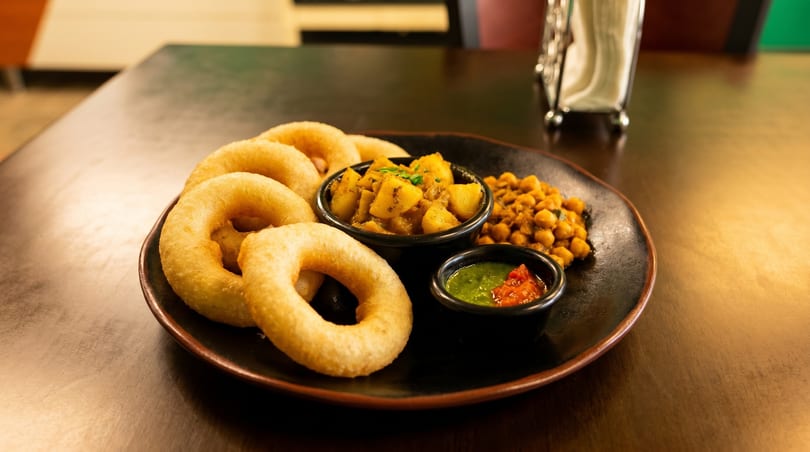 A black plate holds golden fried rings, potato curry, chickpeas, and two dipping sauces.